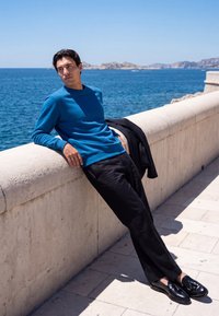 Man in blue sweater and black pants leaning against stone railing by sea on sunny day with distant rocky islands.