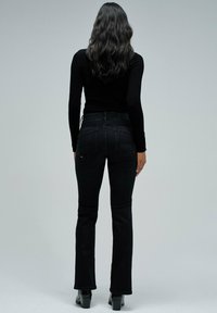 Mujer con cabello largo y oscuro, vistiendo una blusa negra de manga larga, jeans negros de tiro alto y botines negros, de pie contra un fondo sencillo.