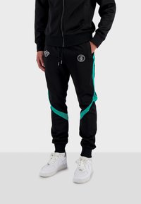 Černé tepláky s tyrkysovými postranními pruhy a vyšívanými detaily, s elastickým pasem a manžetami, v kombinaci s bílými teniskami.