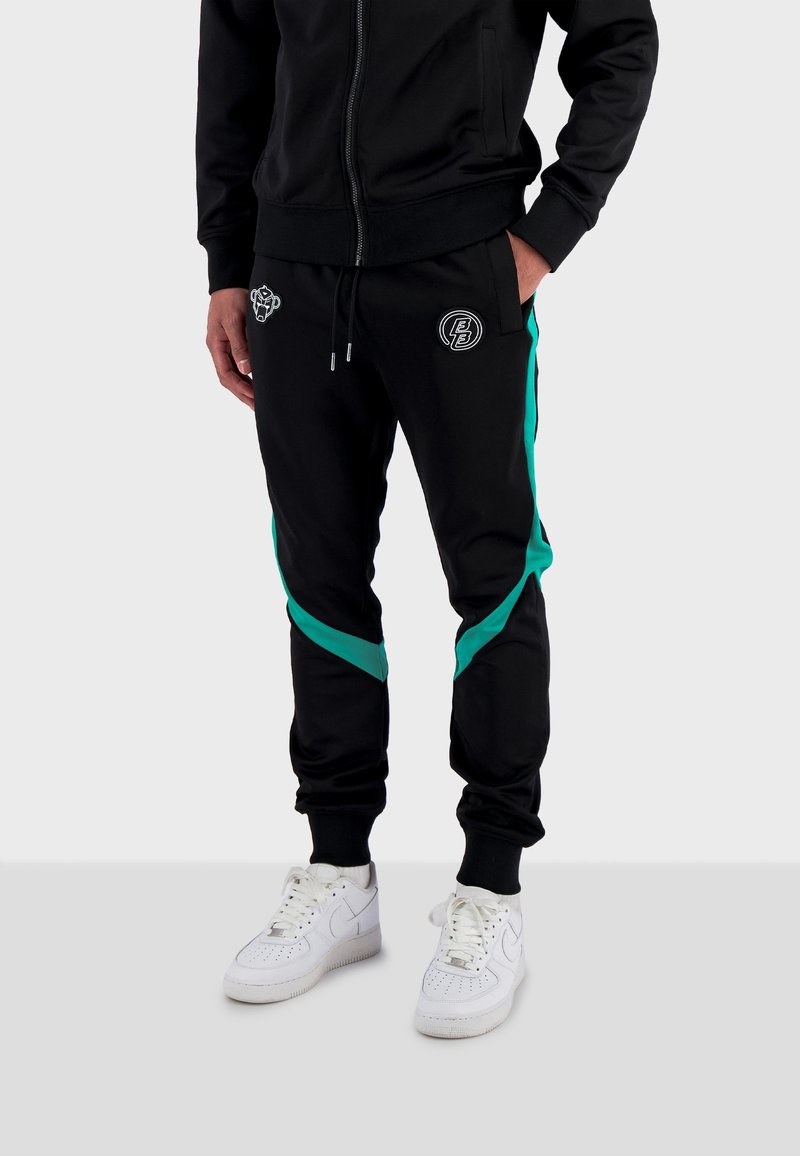 Černé tepláky s tyrkysovými postranními pruhy a vyšívanými detaily, s elastickým pasem a manžetami, v kombinaci s bílými teniskami.