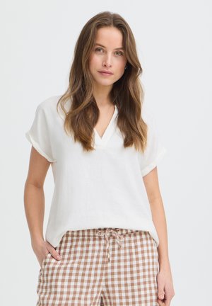 Jeune femme aux longs cheveux bruns portant un chemisier blanc à manches courtes et un pantalon à carreaux marron et blanc, les mains dans les poches.