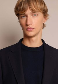 Blazer azul marino sobre un suéter de punto azul oscuro, con un corte entallado, solapas de muesca y una tela texturizada. Líneas limpias y un detalle mínimo.