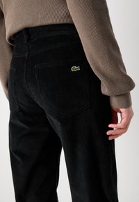 Black corduroy pants with a ribbed texture, featuring a green embroidered logo on the back pocket. Styled with a loose fit and mid-rise waist.