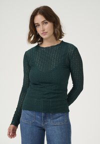 Un haut à manches longues vert foncé en dentelle avec un motif texturé, doté d'un col rond et d'une silhouette ajustée, associé à un jean bleu.