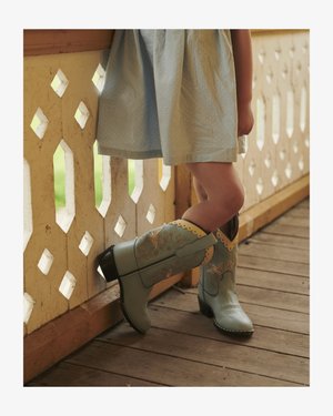 Gyermek világoskék hímzett cowboy csizmát és halványkék ruhát viselve áll egy fa teraszon, díszes korláttal.