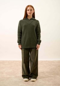 Ciemnozielony sweter na zamek z półokrągłym kołnierzem, o prążkowanej strukturze i kształcie w literę V. Połączony z szerokimi zielonymi spodniami sztruksowymi i białymi butami.