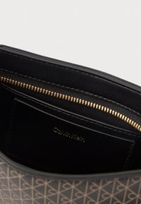 Μαύρη τσάντα χειρός με γεωμετρικό σχέδιο, χρυσό φερμουάρ και λείο δερμάτινο εσωτερικό. Διαθέτει μια μικρή τσέπη με επωνυμία.