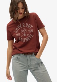 Camiseta color óxido con un diseño gráfico circular en gris claro, combinada con jeans a rayas azules y blancas y un cinturón trenzado.