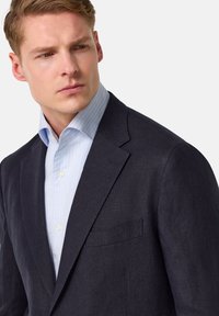 Blazer texturé bleu marine avec un revers à cran; porté sur une chemise rayée bleu clair. Le blazer présente une poche unique sur la poitrine gauche.