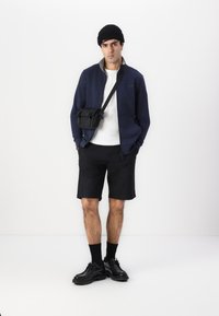Chaqueta navy de cremallera, camiseta blanca, pantalones cortos negros y zapatos negros. Modelo con gorro negro y bolsa cruzada, todo de diseño minimalista.