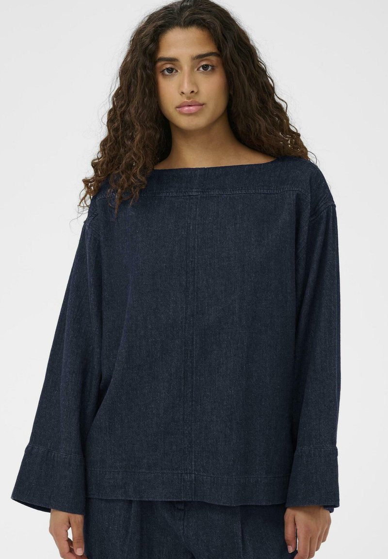 Mladá žena s dlhými kučeravými vlasmi, oblečená v voľnej tmavej rifľovej blúzke s dlhým rukávom a zladených nohaviciach, stojaca pred jednoduchým pozadím.