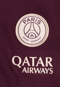 Maglia sportiva borgogna con logo circolare ricamato del Paris Saint-Germain con dettagli beige e "QATAR AIRWAYS" in grassetto beige sottostante.