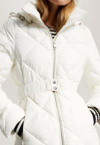 Chaqueta de plumas blanca acolchada con cierre de cremallera, diseño con cinturón y puños a rayas en contraste. Cuenta con una capucha desmontable y botones brillantes.