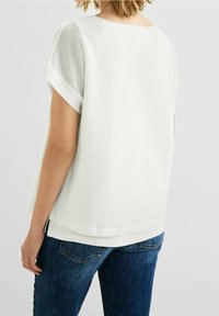 Vit kortärmad topp med en texturerad, öppen vävstruktur. Har en rund halsringning och en lager-på-lager fåll, tillsammans med mörka jeans.