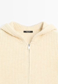 Felpa con zip di colore crema, in maglia a coste. Presenta una texture morbida e un cappuccio ampio per ulteriore calore. Etichetta visibile all'interno del collo.