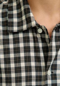 Schwarz-weißer karierten Hemd mit Kragen, das kleine Quadrate mit braunen Nähten umrandet hat, und einem weißen Knopf am Hals.