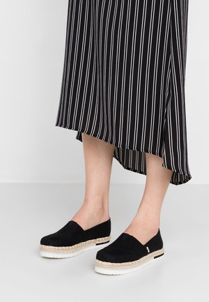 Czarne zamszowe espadryle z beżowymi sznurkami i białymi gumowymi podeszwami, noszone z midi spódnicą w czarno-białe paski.