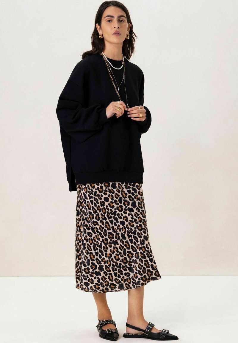 Sweat-shirt noir oversize, jupe midi imprimé léopard, chaussures plates noires. Colliers superposés et boucles d'oreilles dorées. Textures de tissus douces.