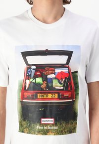 Balta marškinėliai su grafika, kurioje pavaizduota atidaryta automobilio bagažinė, pilna spalvingų daiktų, batų ir licencijos numerio "HNTR 22".
