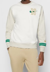 Krämsfärgad och beige sweatshirt med gröna och gyllene detaljer. Har "N"-emblem och Nike-logotyp på vänster bröst och ärmsluten. Tillverkad av bomullsblandning.
