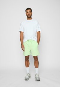 Lyse grønne shorts med sidelommer og snørelukning, parret med en løs hvid t-shirt og grå træningssko.