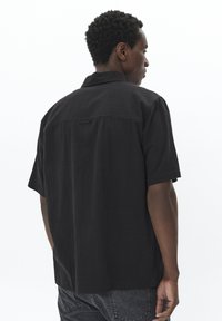 Chemise noire à manches courtes avec une coupe décontractée, tissu texturé et un détail en boucle au col arrière. Associée à un jean en denim foncé.