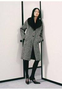 Cappotto a scacchi grigi con colletto in pelliccia sintetica nera, caratterizzato da due bottoni e tasche laterali. Indossato sopra leggings neri e tacchi a punta.