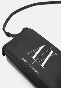 Sort rektangulær taske med en struktureret overflade, der har et fremtrædende hvidt "AX" logo og teksten "ARMANI EXCHANGE". Inkluderer en aftagelig rem.