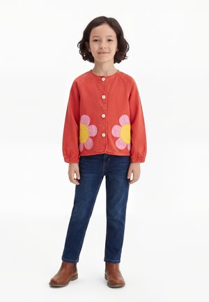 Dítě s krátkými tmavými vlasy má na sobě červenou bundu s růžovými a žlutými květinovými nášivkami, modré džíny a hnědé boty, stojí před bílým pozadím.