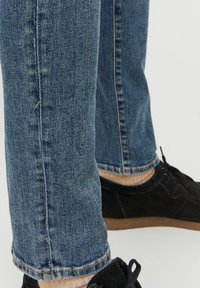 Jeans en denim bleu à coupe slim avec une texture décolorée, des coutures surpiquées et un ourlet roulé. Portés avec des chaussures en suède noires et des chaussettes de couleur claire.