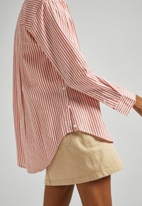 Camicia a righe rossa e bianca con maniche lunghe, caratterizzata da un orlo arrotondato e bottoni visibili di lato. Indossata con pantaloni corti color khaki.