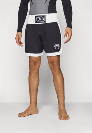 Hombre con pantalones cortos de boxeo Venum Pro en blanco y negro, de pie descalzo con las manos relajadas a los lados contra un fondo liso.