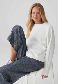 White, long-sleeve sweater with a relaxed fit and crew neckline, paired with dark, textured wide-leg trousers and white sneakers.
