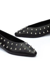 Chaussures plates noires à bouts pointus ornées de clous ronds en argent répartis uniformément sur la surface supérieure, sur fond blanc.