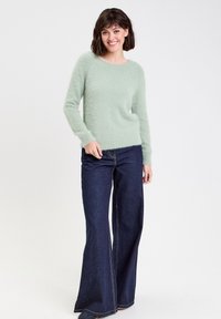 Hellgrüner, flauschiger Pullover mit Rundhalsausschnitt und langen Ärmeln, kombiniert mit dunkelblauen Schlagjeans, die sichtbare Nähte aufweisen.