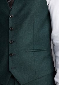 Gilet boutonné vert foncé porté sur une chemise blanche, montrant cinq boutons noirs et une poche passepoilée à l'avant.