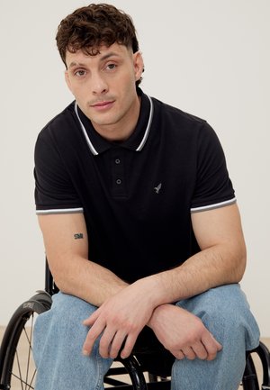 Άνδρας με κοντά σγουρά μαλλιά, φοράει μαύρο πόλο και τζιν, κάθεται σε αναπηρικό καροτσάκι, κοιτάει μπροστά με τα χέρια σταυρωμένα.