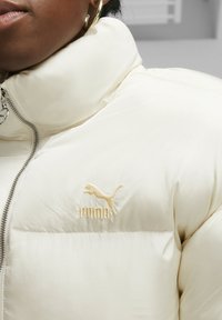 Cremefarbene Pufferjacke mit hohem Kragen, ausgestattet mit einem Reißverschluss und einem leicht goldenen, bestickten Puma-Logo. Glatter, matter Stoff.