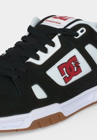 Ασπρόμαυρο αθλητικό παπούτσι με κόκκινο λογότυπο DC, μαύρα κορδόνια και σόλα χρώματος καουτσούκ, που δείχνει λεπτομέρειες από το πλάι και μπροστά.