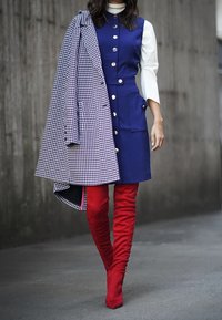 Žena m�á na sobě bezrukávové modré šaty na knoflíky, bílé dlouhé tričko, červené nadkolenky, a nese modro-bíle kostkovaný kabát naruby na rameni.