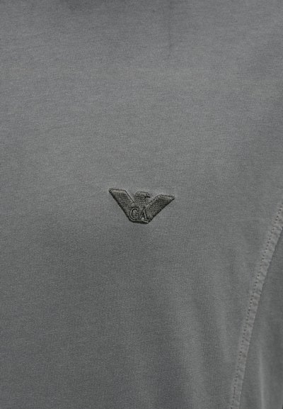 Šedé tričko s krátkým rukávem z měkké látky s reliéfním logem orla a nápisem "GA" na hrudi. Celkově hladká textura.