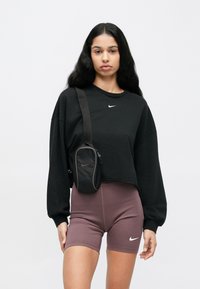 Μαύρο κοντό φούτερ με το λογότυπο της Nike, σετάρεται με ψηλόμεσο κοντό ποδηλατικό σορτς σε αποχρώσεις μωβ. Μικρή μαύρη τσάντα ώμου με φερμουάρ και λογότυπο.