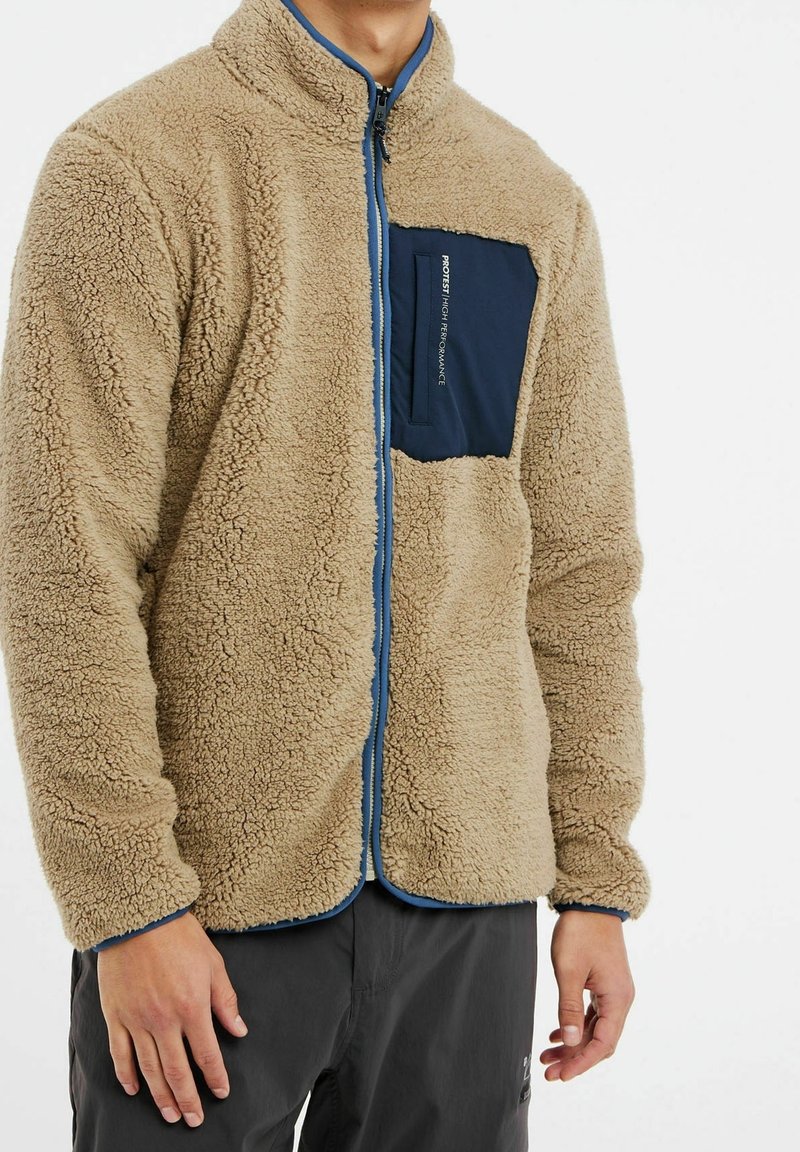 Homme portant une veste en polaire sherpa beige avec une fermeture éclair bleue et une poche poitrine bleu marine sur un pantalon foncé.