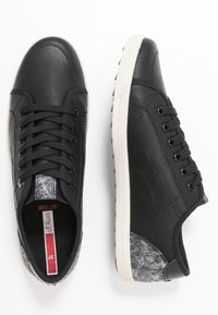 Black sneakers with a textured upper, featuring a silver accent at the heel and a white rubber sole with tread. Laced closure design.