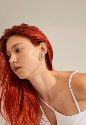 Strieborné náušnice majú textúrovaný dizajn s lesklým akcentom. Modelka má dlhé, rovné, červenohnedé vlasy a nosí biele tričko.