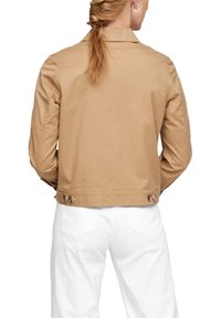 Osoba z długimi włosami w niskim kucyku nosi beżową kurtkę zapinaną na guziki i białe spodnie, przedstawiona z tyłu na białym tle.