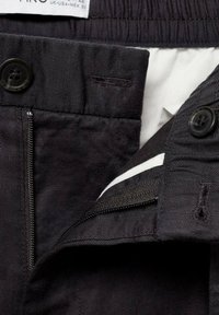 Black pants with a zip fly, button closure, and elastic waistband; features a white inner lining and a label with size information.