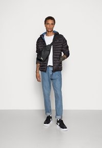 Svart pufferjakke med hette, blå fôr, hvit t-skjorte, lyseblå jeans og svarte sneakers. Svart crossover-veske med logo.
