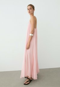 Woman wearing a long, flowing pink dress and black sandals stands sideways against a plain white wall, with a silver bracelet on her arm.