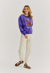 Fialová mikina s grafikou hnědého pudla a textem "Objímání, Kmitání, Pudlové Směšky." V doplnění s krémovými kalhotami a botami s leopardím vzorem.